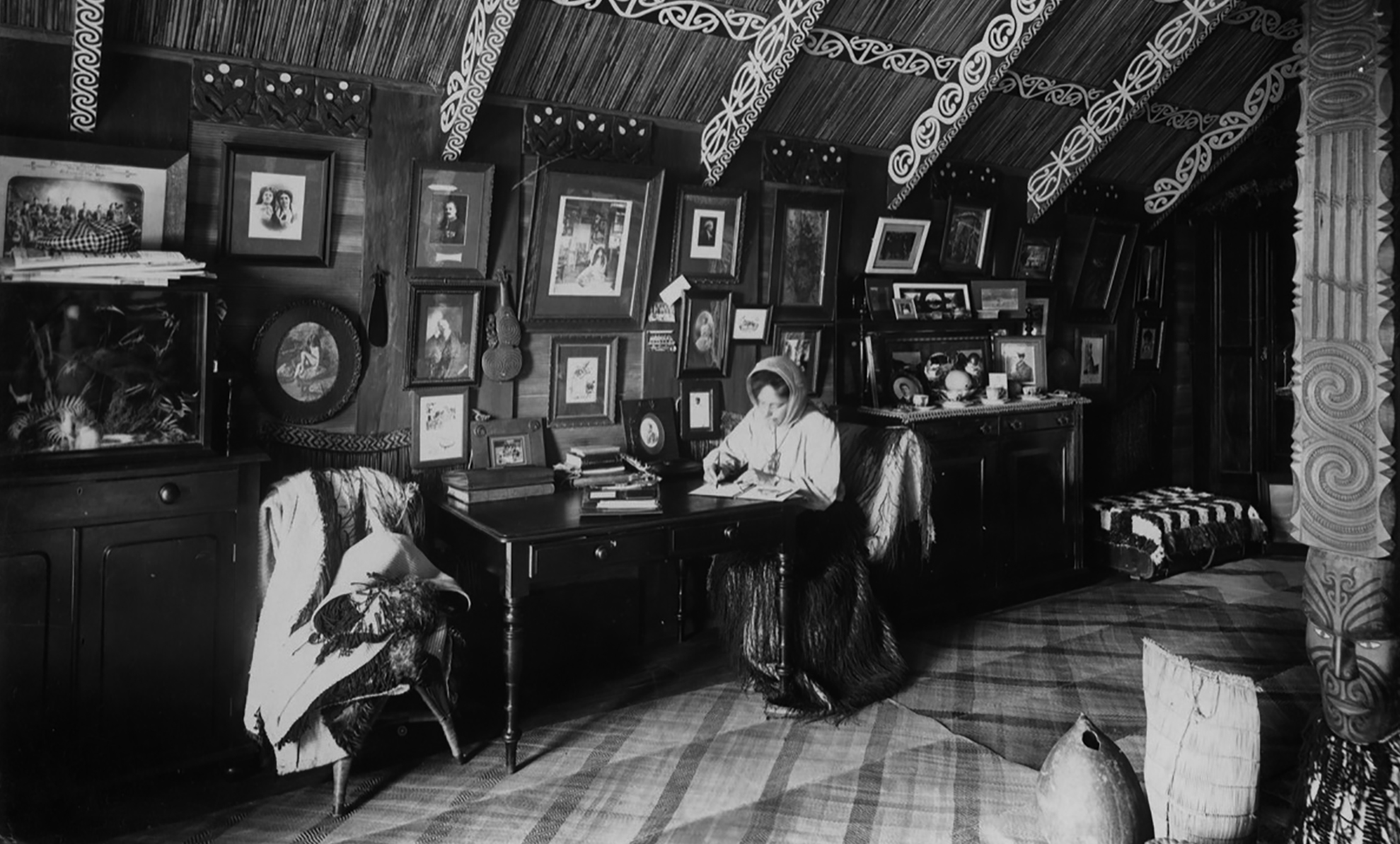 Interior view of a Maori house with a person sat at small table against a wall with many framed pictures.