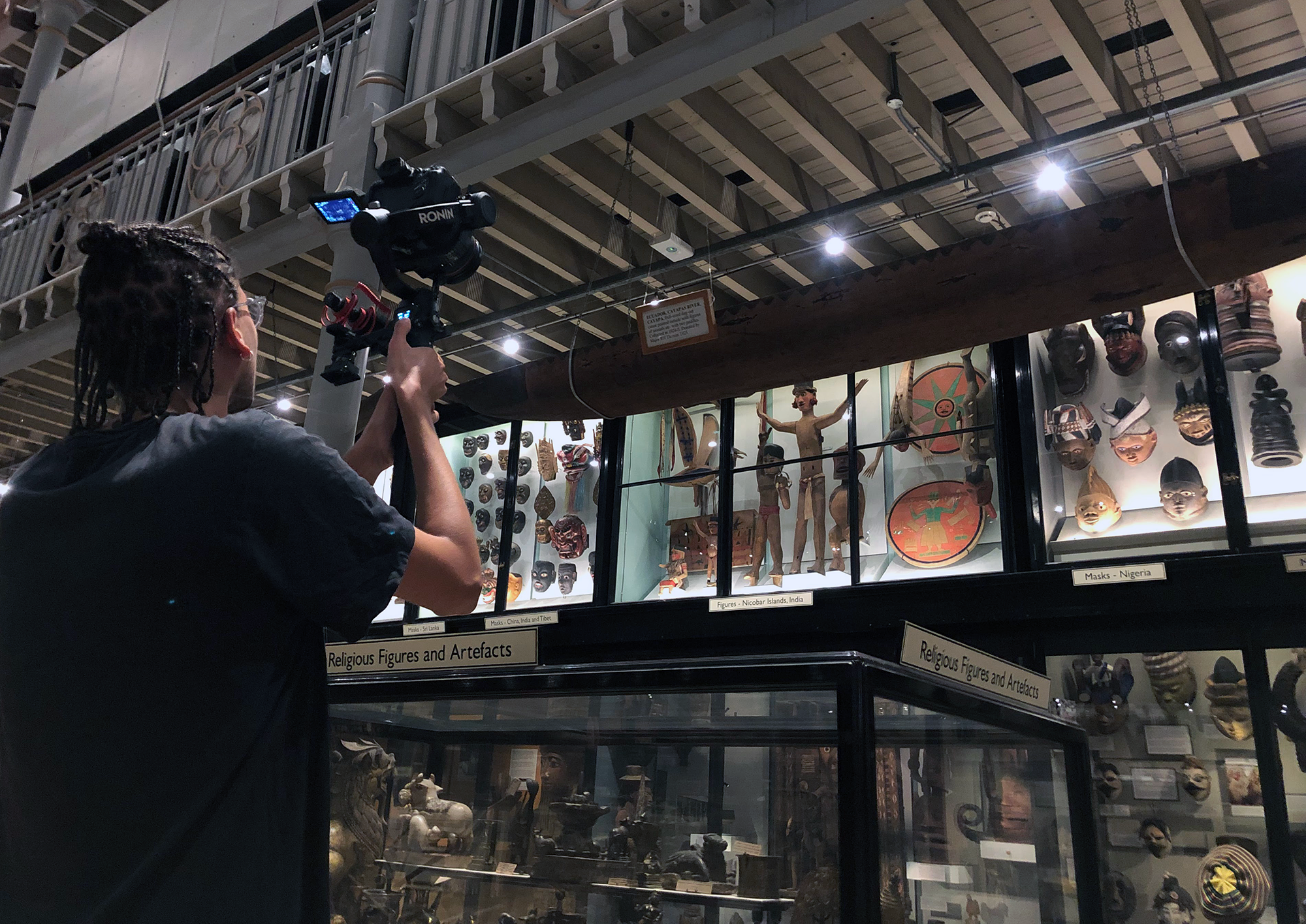 a person lifts a digital camera on a gimble above their shoulders, pointed at objects display in tall glass cases at the museum.