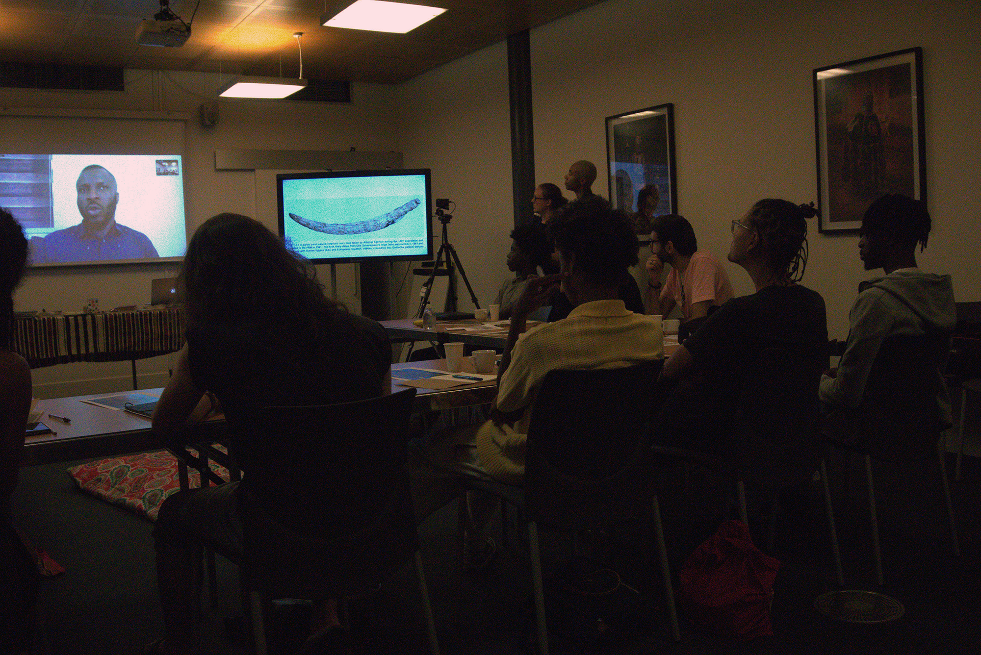 a group of people face two screens in a darkened lecture room, one showing a speaker giving an online talk and another showing a carved ivory tusk