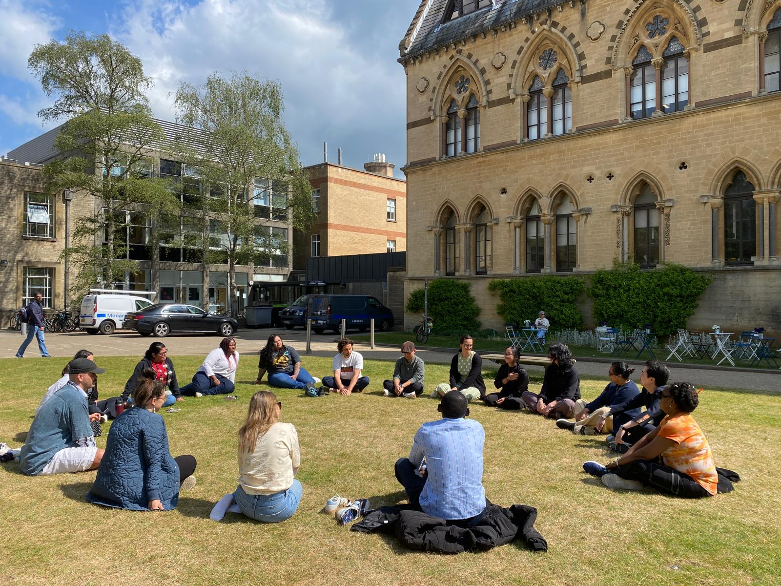 A group of people sit in a circle facing each other on a grassy lawn