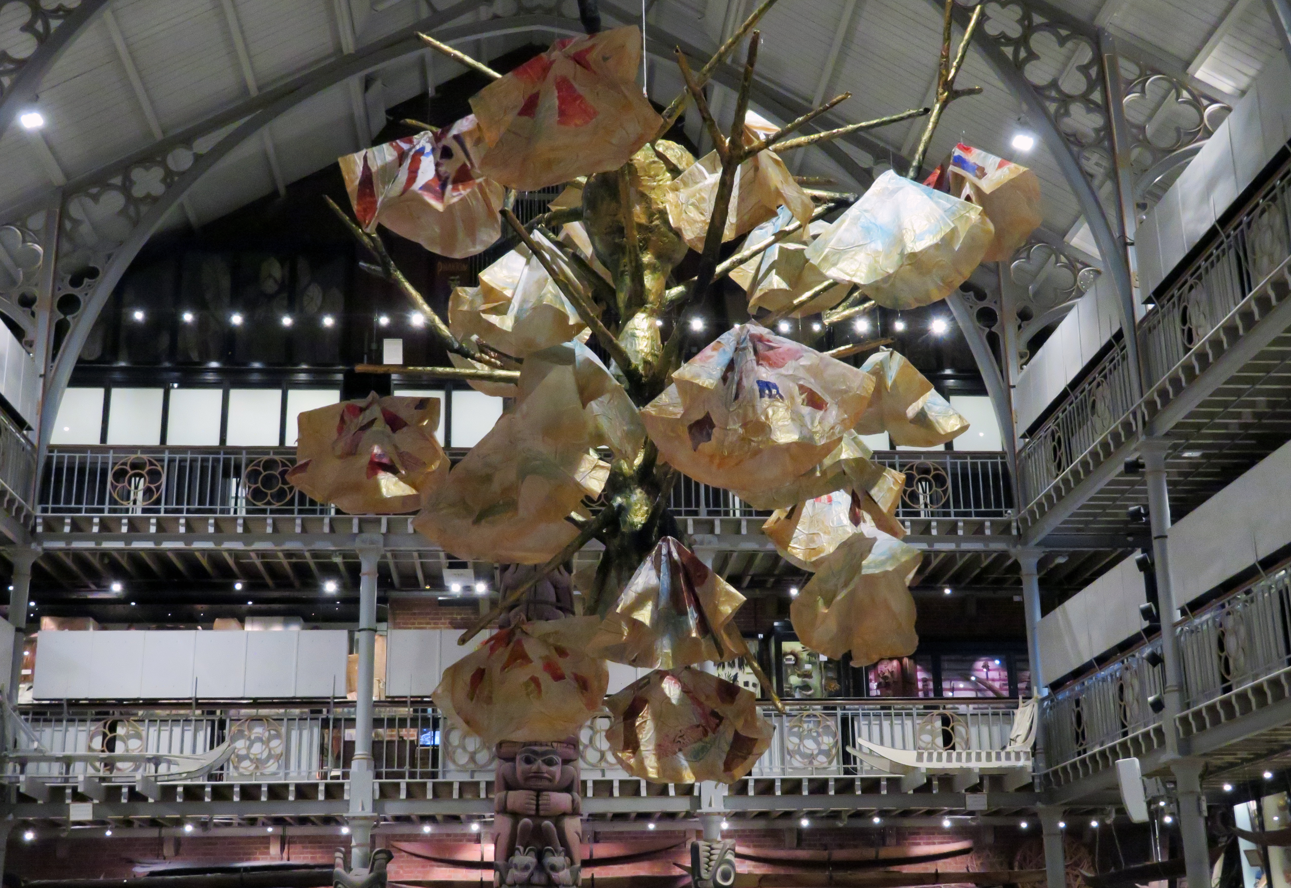 Close up view of a branching paper sculpture in front of a backdrop showing the roof and railings of gallery floors of a museum.
