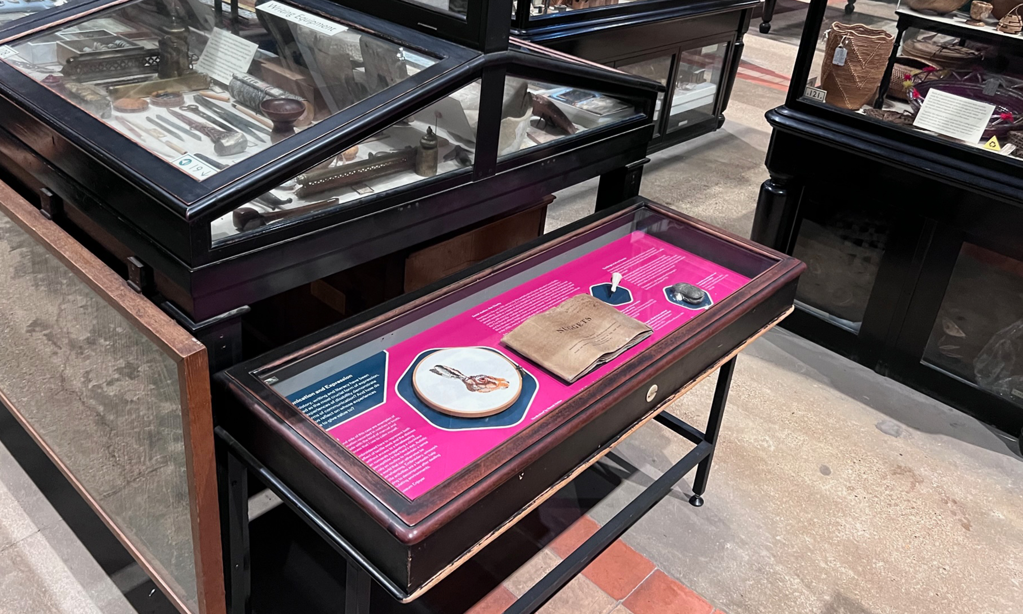 View of a desktop display case with bright pink graphic board in a museum full of tightly packed display cases full of objects