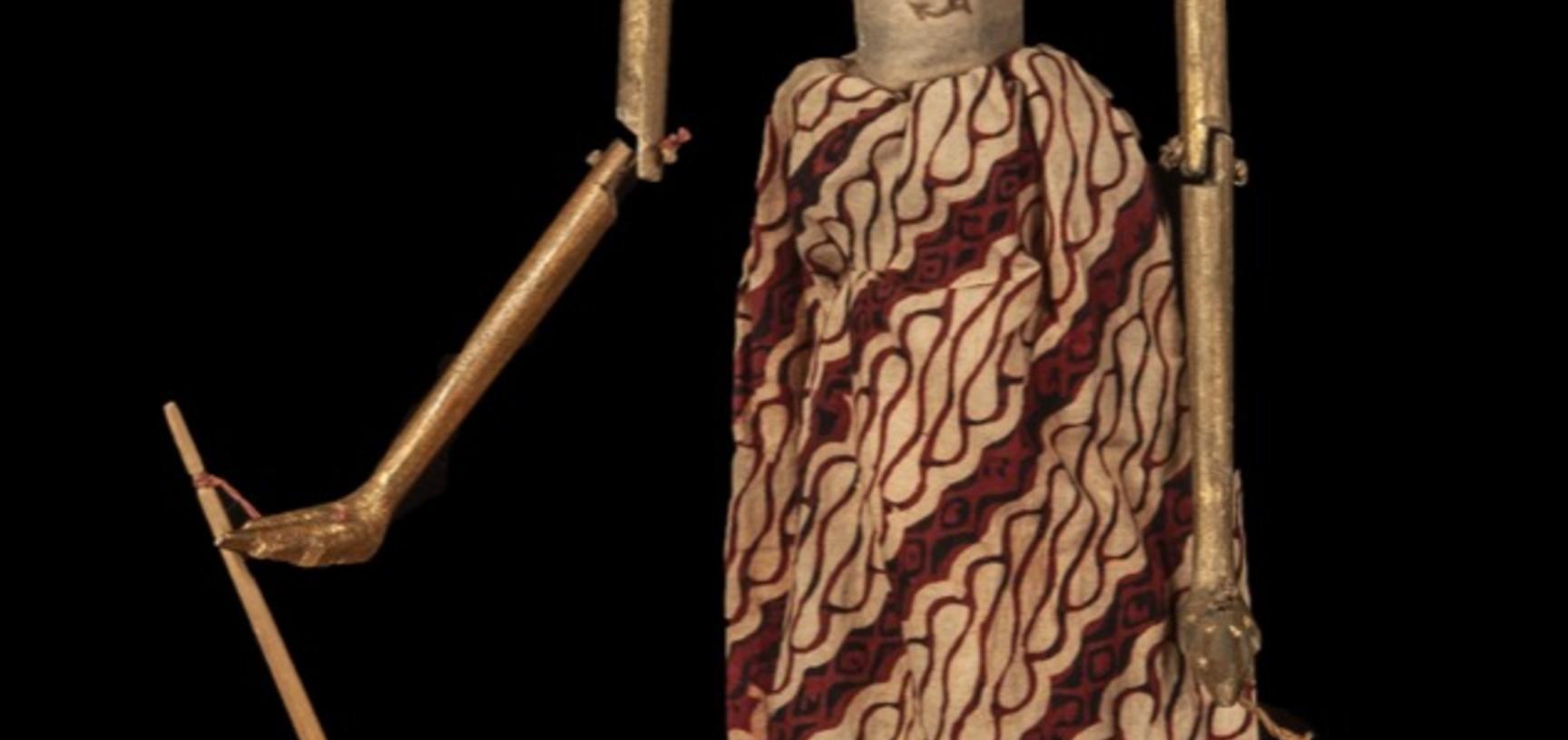 Wooden puppet with white face, body and hair painted gold, with long skirt of red and brown patterned cloth.