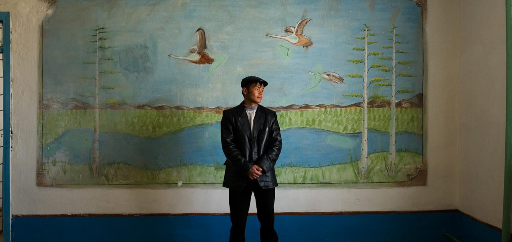 A student posing for a portrait in front of a mural inside his school in Muynaq. This city has been devastated by the shrinking of the Aral Sea. Muynaq, Uzbekistan. Photograph by Carolyn Drake. March 2008.