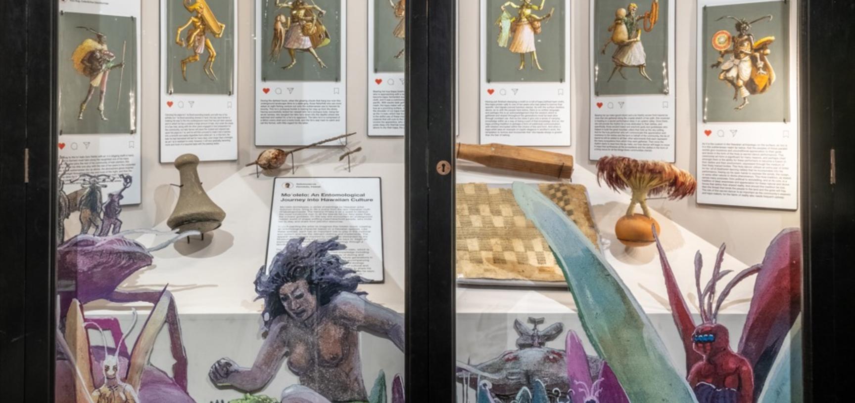 Case display showing seven watercolour illustrations alongside objects featured in the paintings, with colourful cut outs of illustrated figures framing the glass front.