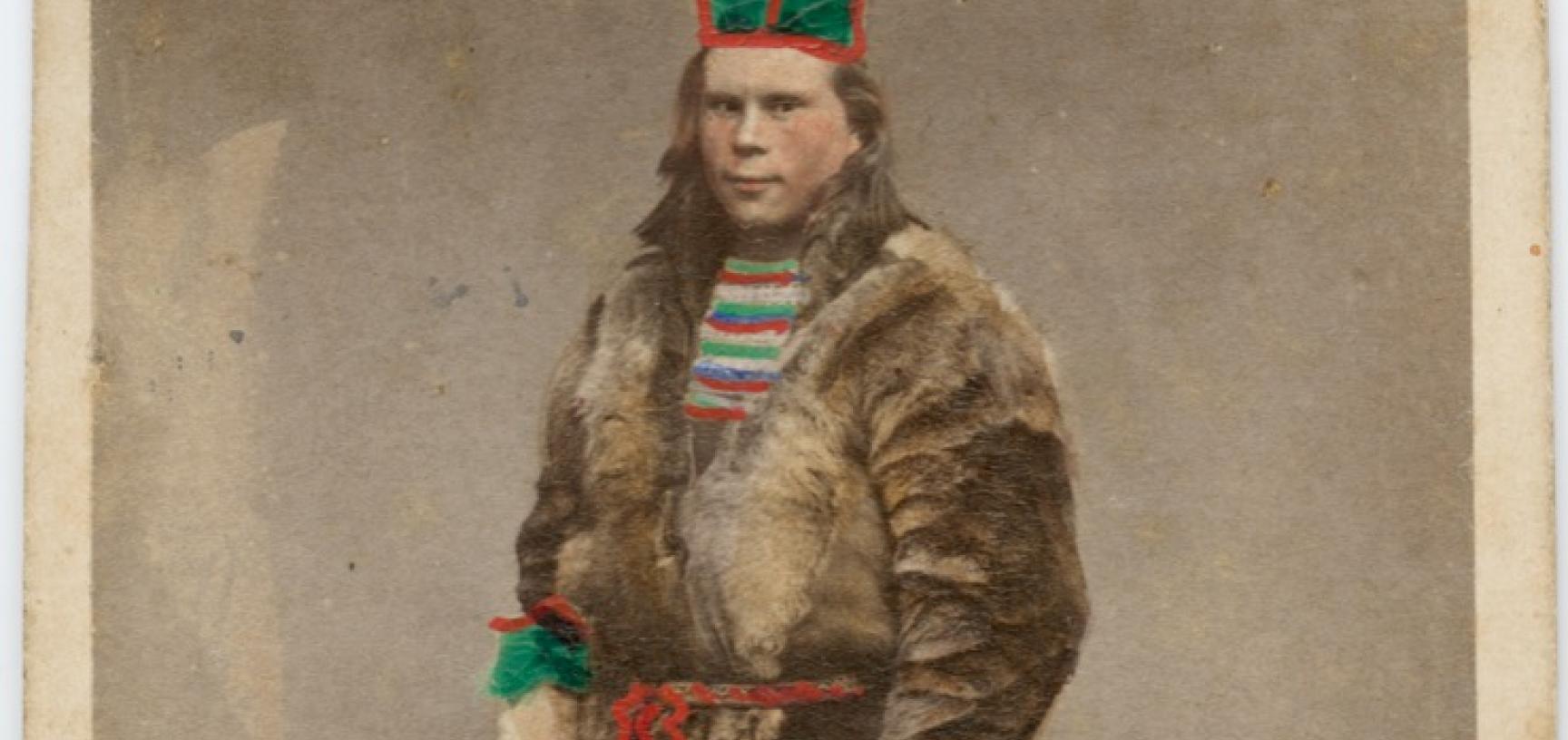 Hand-coloured studio portrait of a Saami man, standing, wearing an animal skin jacket and a distinctive hat, identified in a handwritten note as being from Luleå, Sweden. Photograph by the Rosalie Sjöman studio. Stockholm, Sweden. Circa 1870s. (Copyright 
