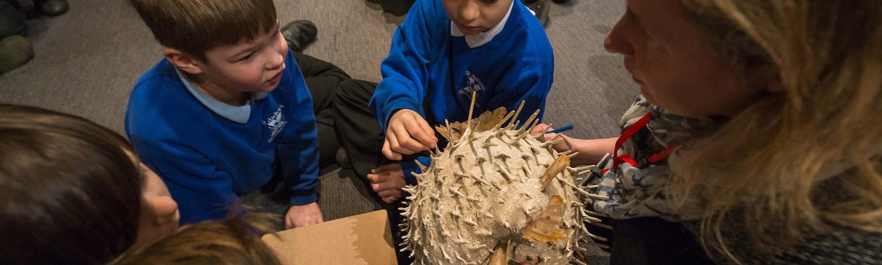 Primary school pupils sit on floor and handle an ivory coloured, oval-shaped helmet with long spikes protruding from all around it