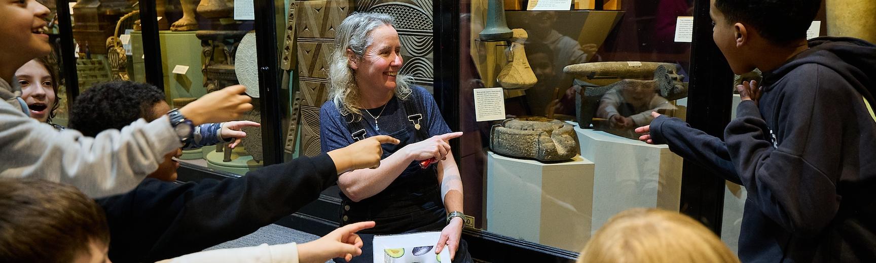Education officer and school group sitting on floor pointing at objects in museum display case.
