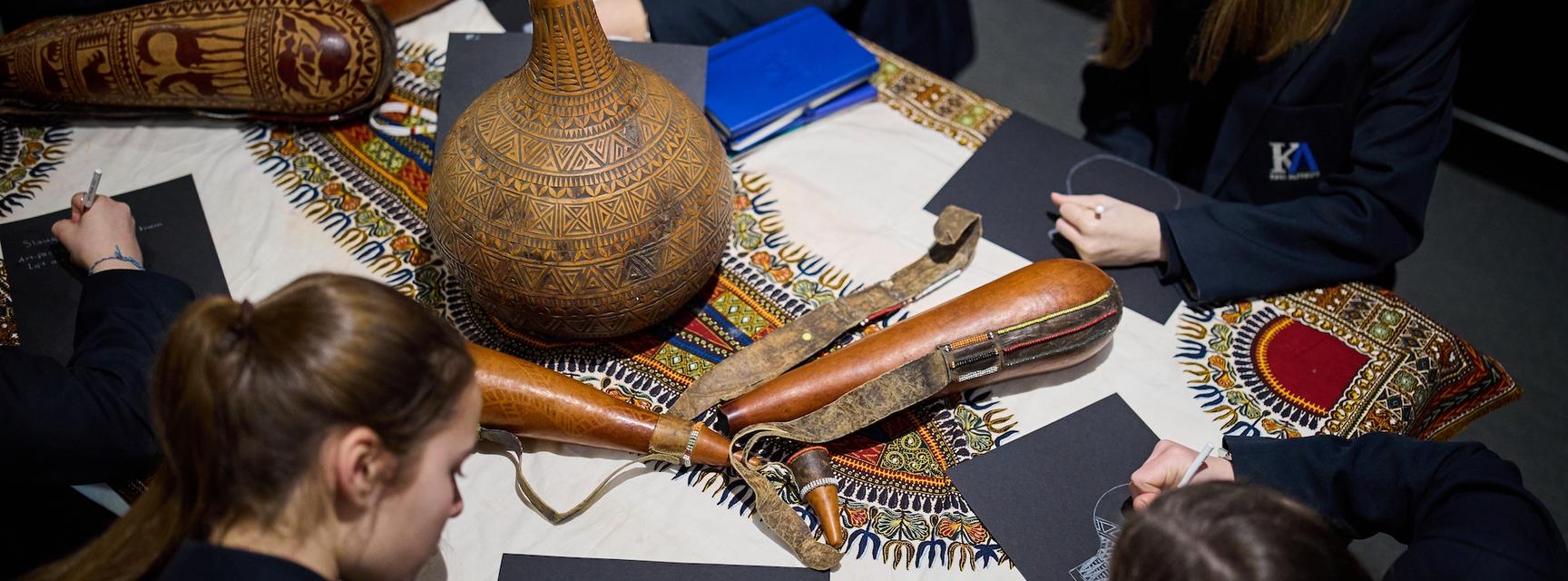 School students sitting around table drawing decorative gourds and textiles which are grouped on the table.