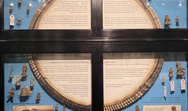 Photograph of a shallow display case showing a graphic reproduction of a large circular ungo (basket) surrounded by lots of small clay figures, photographs of people and text labels. 