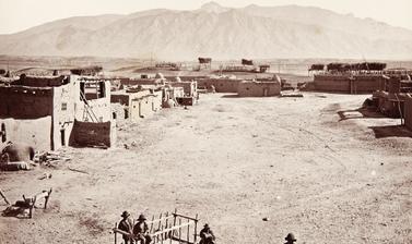 View of the main plaza of Sandia Pueblo, with a wooden cart (known as a carreta) and several people prominent in the foreground, and the Sandia mountains in the distance. 