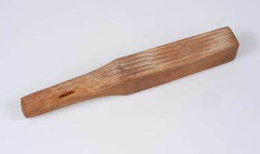 A rectangular block of wood used for beating bark cloth. It has lines engraved at one end and narrows slightly at the other end to form a handle.