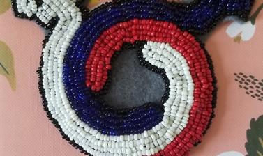 Red, white and blue beads forming a circular shape with a cross projecting from the base and an arrow projecting from each side at the top. Placed on a floral background. 