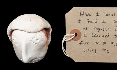 White clay formed into a face with a rectangular brown paper handwritten luggage tag style label