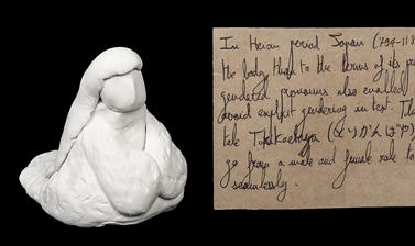 White clay formed into a seated figure with a rectangular brown paper handwritten luggage tag style label