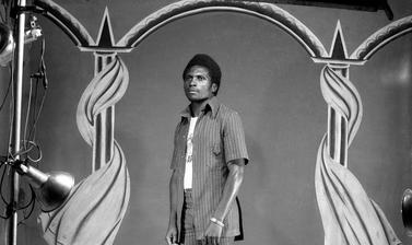 Man dressed in contemporary fashion standing in front of Touselle’s third painted backdrop of pillars draped with curtains. Photograph by Jacques Touselle. Mbouda, Cameroon. About 1975.