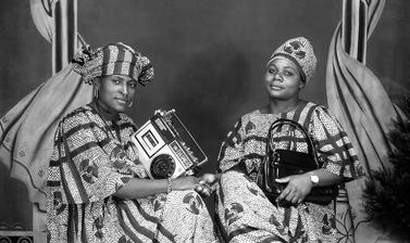 Portrait of two friends holding a cassette player and a handbag, possibly commemorating their purchase of these valued consumer goods. Photograph by Jacques Touselle. Mbouda, Cameroon. About 1975.