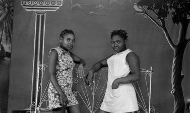 Young women leaning on an ornamental iron grille in front of a painted studio backdrop. Photograph by Jacques Touselle. Mbouda, Cameroon. Early 1970s.