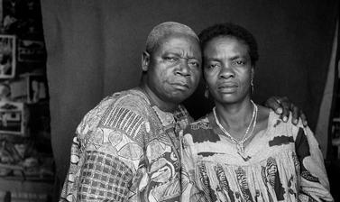 Couple posed for a marriage certificate portrait, with prints on the studio wall visible behind the cloth backdrop. Photograph by Jacques Touselle. Mbouda, Cameroon. Early 1970s.