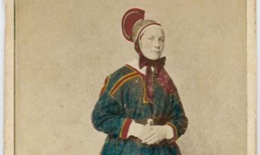 Hand-coloured studio portrait of a Saami woman, identified as a wife (or married woman), standing, wearing a distinctive hat. Photograph by the Marcus Selmer studio. Bergen, Norway. Circa 1873.