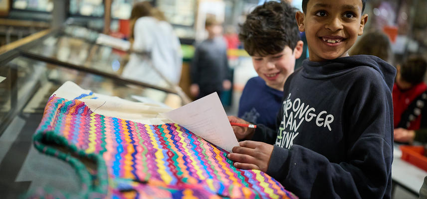 Two boys wearing nave blue tops looking at a multi-coloured striped garment laid out on a case top.