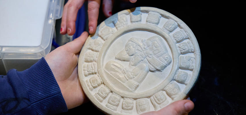 Close up of hands holding a circular white plaque decorated with a central figure and emblems around the edge.