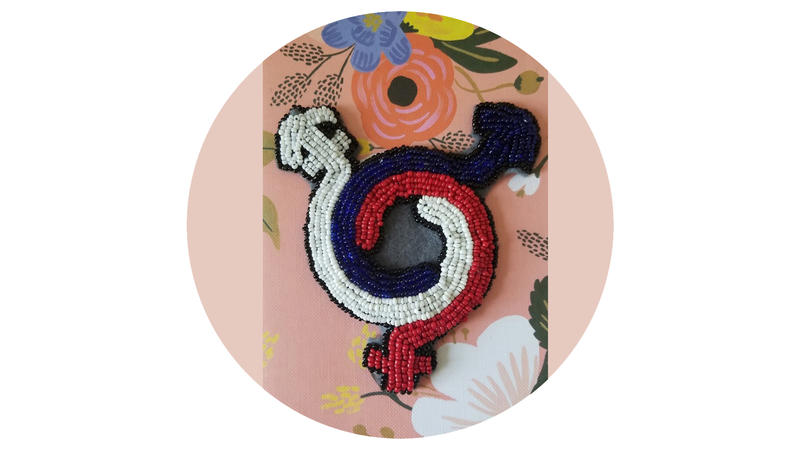 Red, blue and white beads stitched together in three swirling arrows, creating a circular beadwork parth with three protruding arrows, positioned on a pink floral cloth.