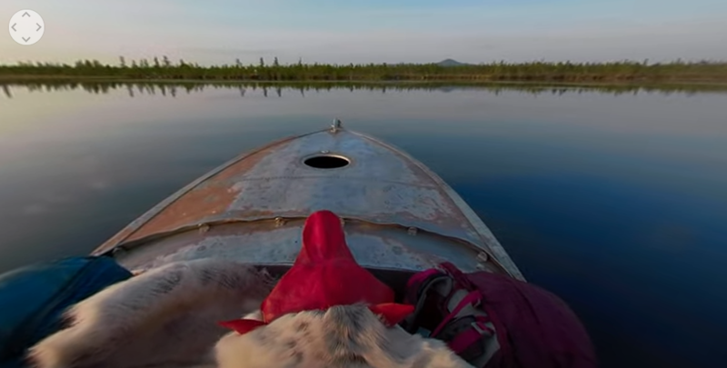 Still from a film showing a red wolf mask on the prow of a small motorboat, looking towards open water of lake.