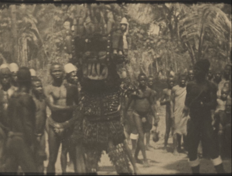 A film still showing a grainy sepia image of a group of people surrounding a person in a masquerade outfit