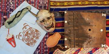 Selection of objects including Japanese mask, beaded animal face and wooden and metal thumb piano laid our on multi-coloured textiles