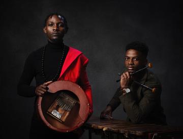Two musicians dressed in black, one with red shawl draped on shoulder, holding an mbira and wooden percussion instrument