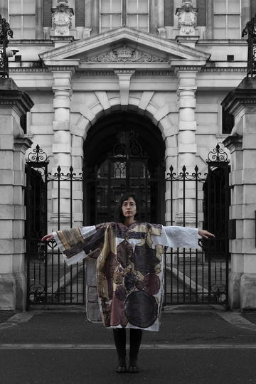 Female standing with arms outstretched in front of a classical stone building, wearing an elaborately embroidered tunic.