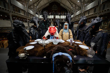 a group of people stand around a table covered with a faux bearskin, some dressed as crow birds, and one as a wolf.