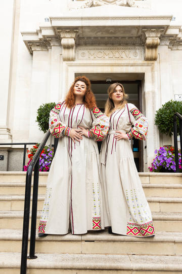 Two women wearing long white dresses with colourful embroidery standing on stone steps