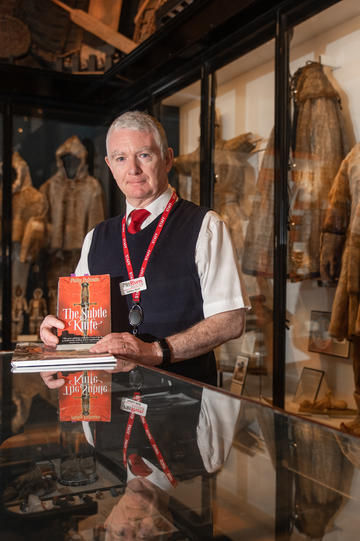 Male member of staff standing by museum display cases holding a copy of His Dark Materials book