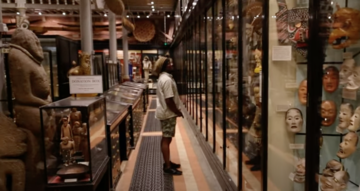 A person stands between glass display cases in a museum looking up at masks on display
