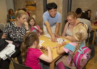 Two women and three children sitting around a table playing games with male museum volunteer assisting.