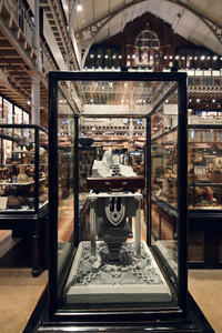 Glass display case containing an intricate sculpture with a miniature landscape emerging from a wooden drawer and mounted on geometric architectural structure.